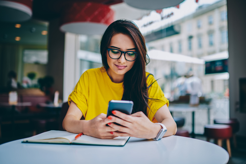 Young woman in spectacles writing a job ad on her mobile device with a notebook in front of her.